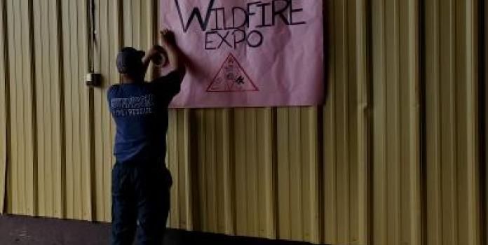 A man hangs a paper sign that says "wildfire expo" on an exterior wall