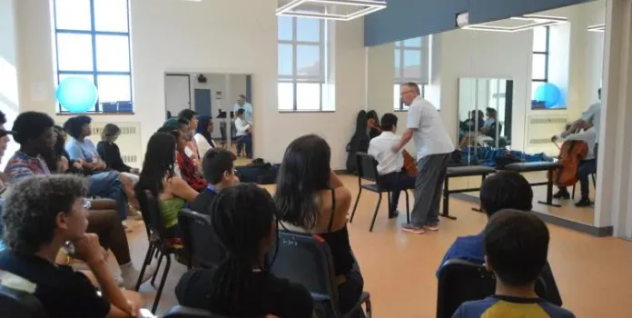 A man instructs young musician in a classroom filled with students