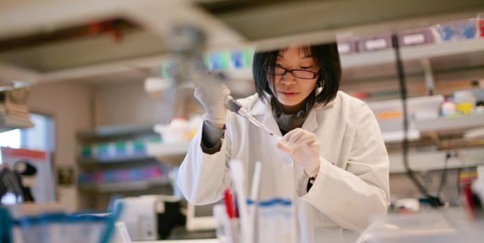 A researcher in a white lab coat and gloves using a pipette to carefully transfer liquid into a small vial in a laboratory setting.