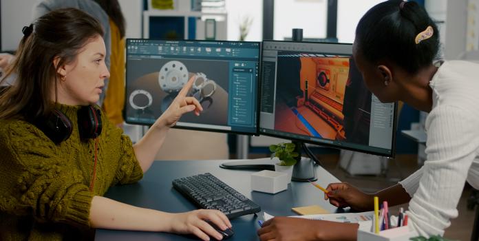 Two designers collaborating at a dual-monitor workstation, discussing game design.