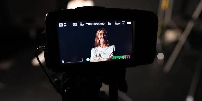 A close-up shot of a professional camera monitor displaying a smiling woman during a video shoot in a dark studio.