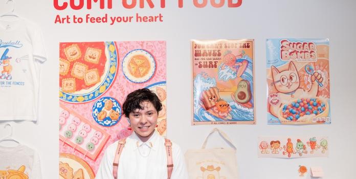 A Denver student in front of their art exhibition wall titled "COMFORT FOOD: Art to feed your heart," featuring vibrant illustrations of food characters on posters, t-shirts, and tote bags.