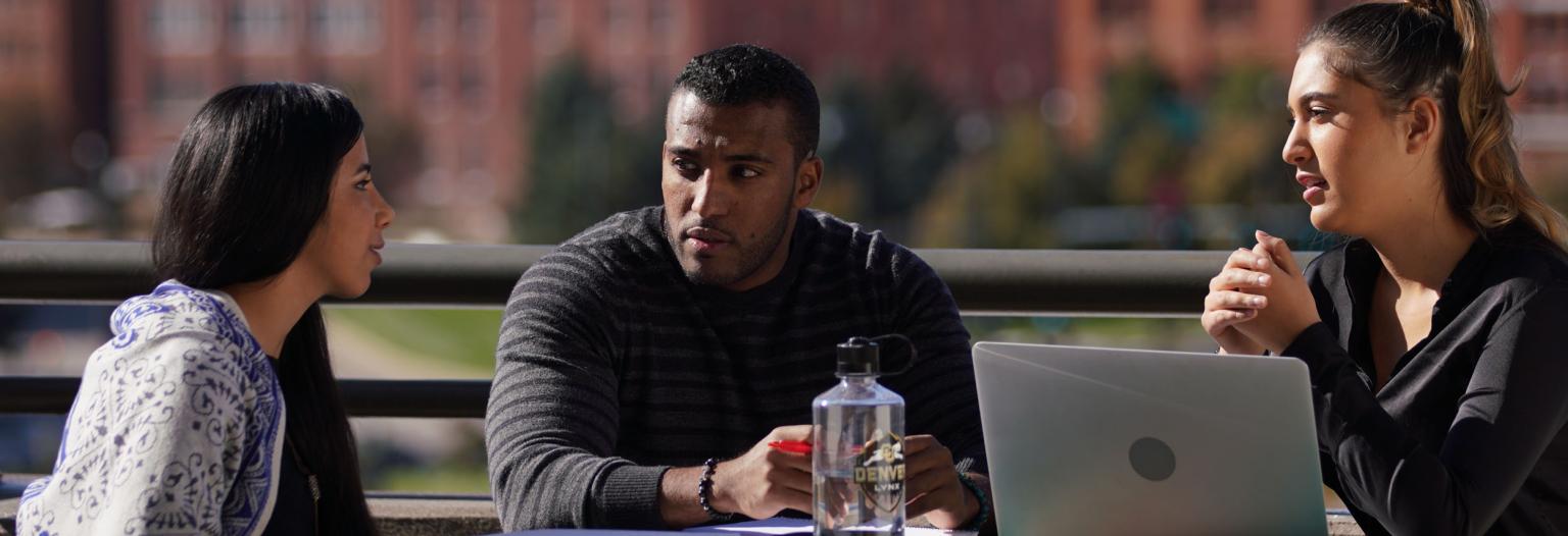 Three students sit around a green outdoor table, having a discussion. On the table are a laptop, notebooks, and a clear water bottle with the CU Denver Lynx logo. 
