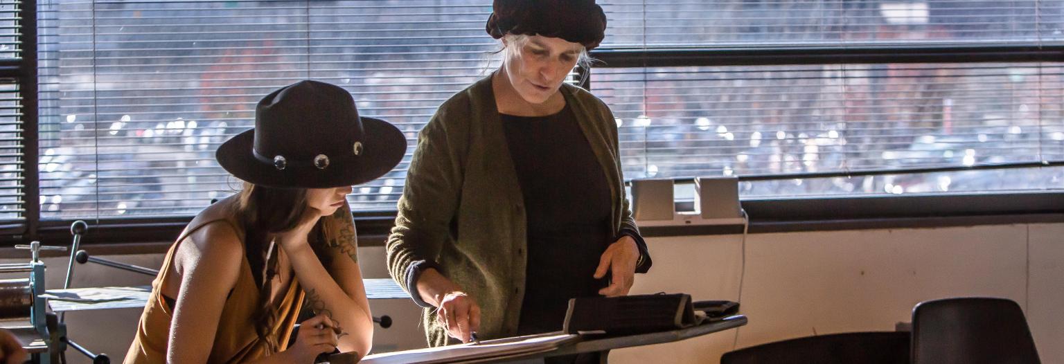 Two women working together at a drafting table in a bright studio with large windows overlooking a city and mountains.